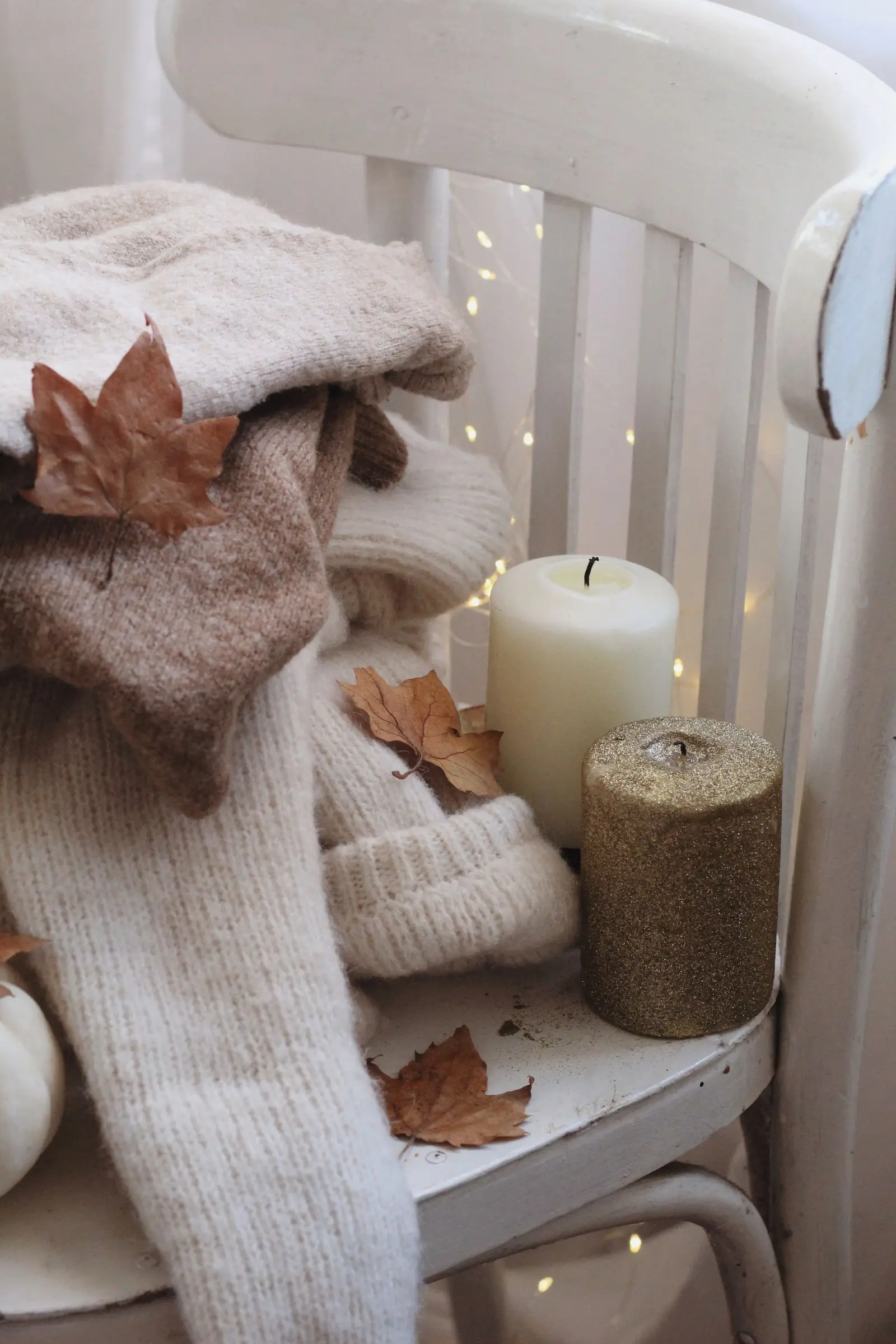 Warm autumn setting featuring folded sweaters, candles, and dried leaves on a white chair.